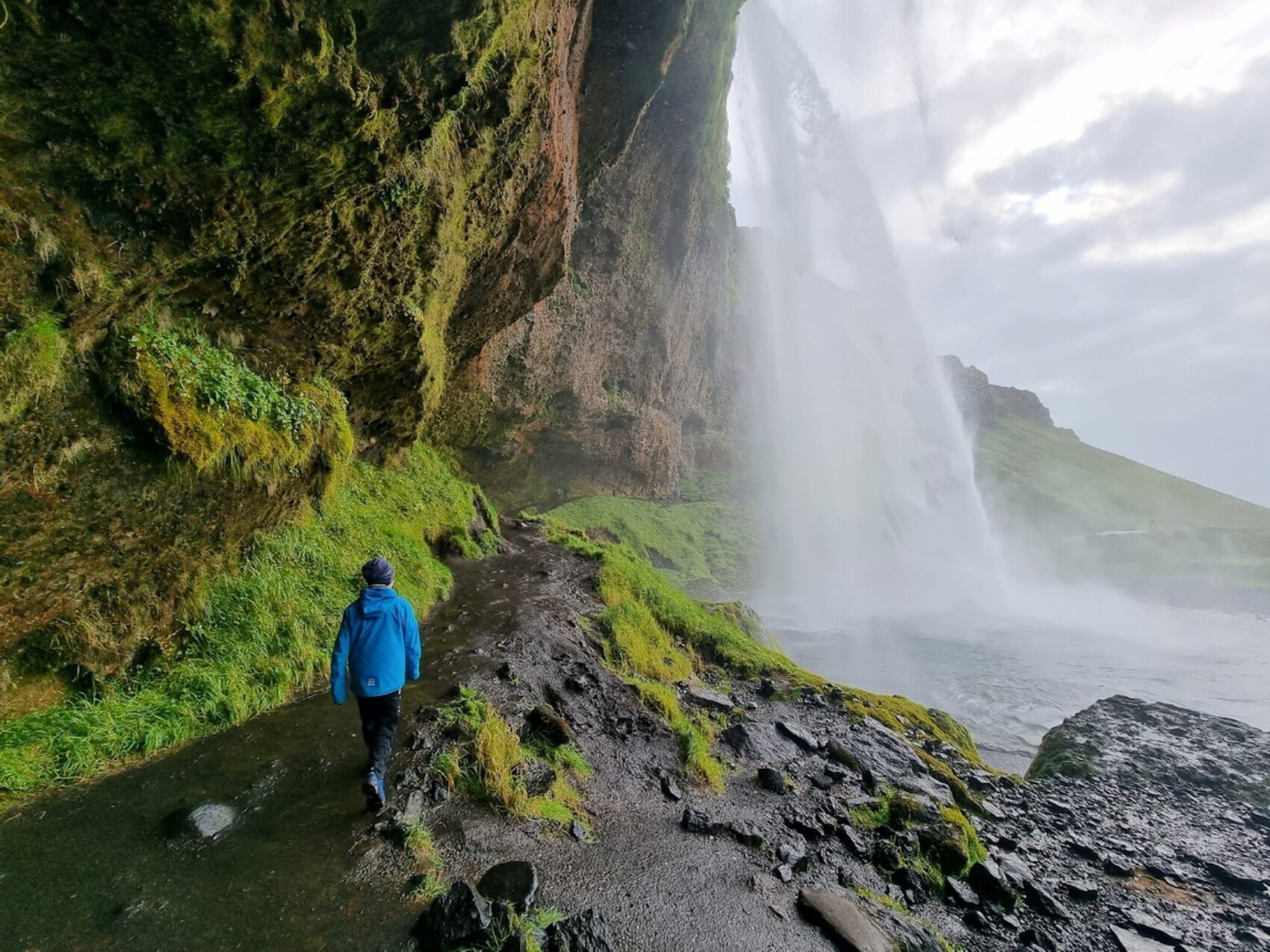 Person läuft hinter Wasserfall auf Trampelpfad entlang