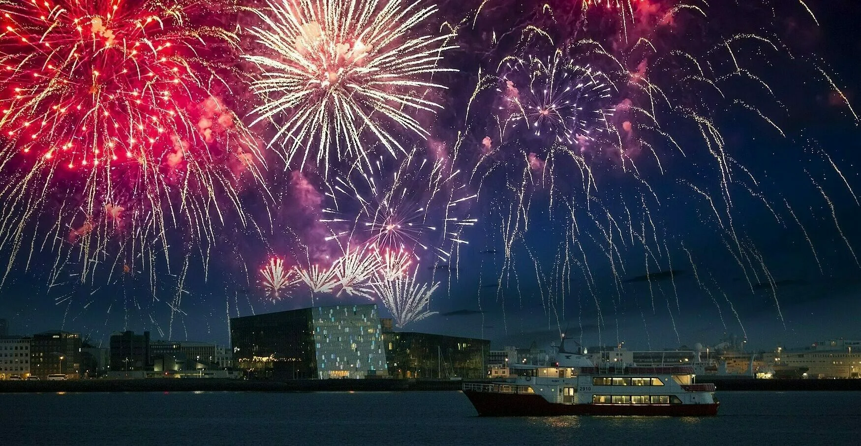 Silvester Feuerwerk über Reykjavik, auf dem Meer ist ein Boot zu sehen, mit der Konzerthalle Harpa im Hintergrund