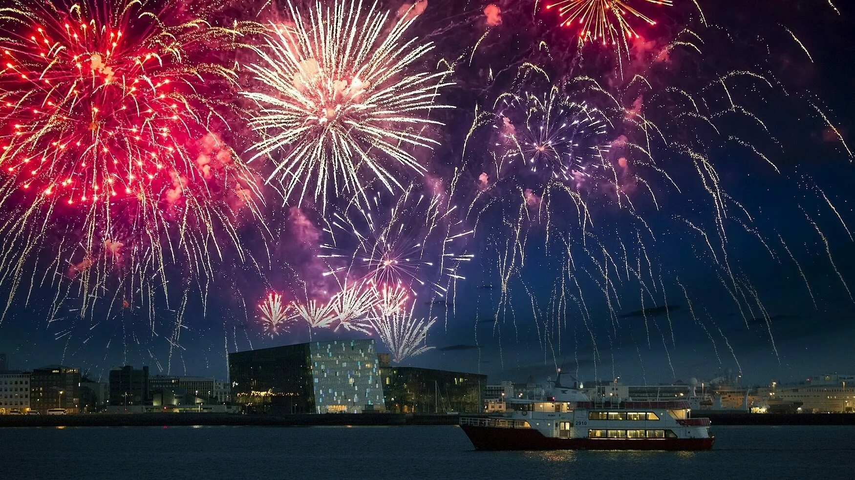 Silvester Feuerwerk über Reykjavik, auf dem Meer ist ein Boot zu sehen, mit der Konzerthalle Harpa im Hintergrund