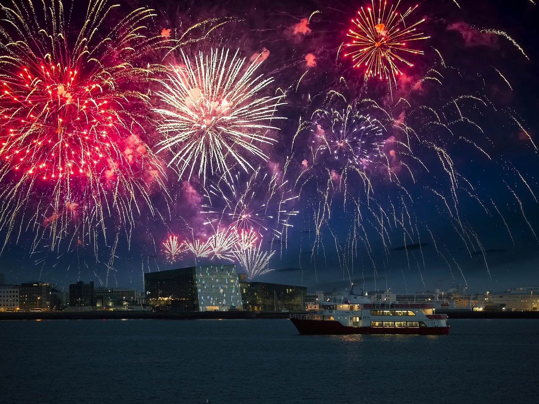 Silvester Feuerwerk über Reykjavik, auf dem Meer ist ein Boot zu sehen, mit der Konzerthalle Harpa im Hintergrund