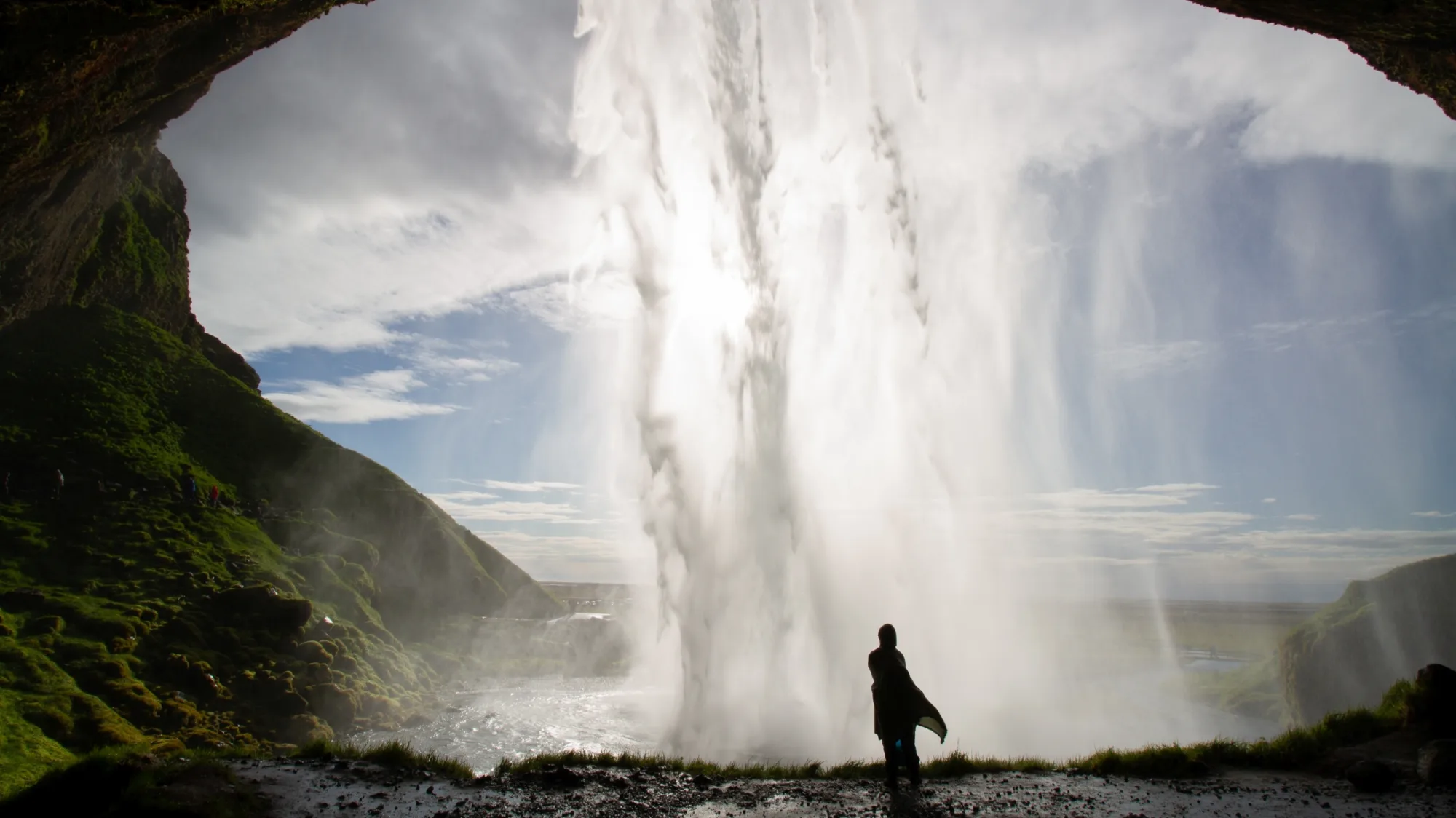 Eine Person steht bei schönem Wetter hinter dem Seljalandsfoss Wasserfall in Island