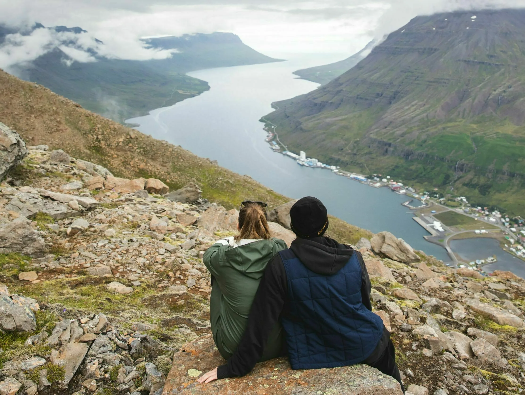 Zwei Wanderer in Ostisland genießen den Ausblick auf den Fjord, sitzen auf einem Felsen mit Rücken zum Bildbetrachter