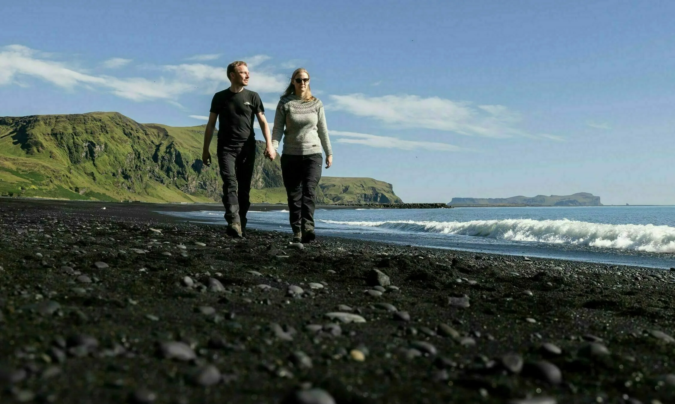 Ein Pärchen welches händchenhaltend an einem schwarzen Strand mit Blick auf Meer und Berge spazieren geht