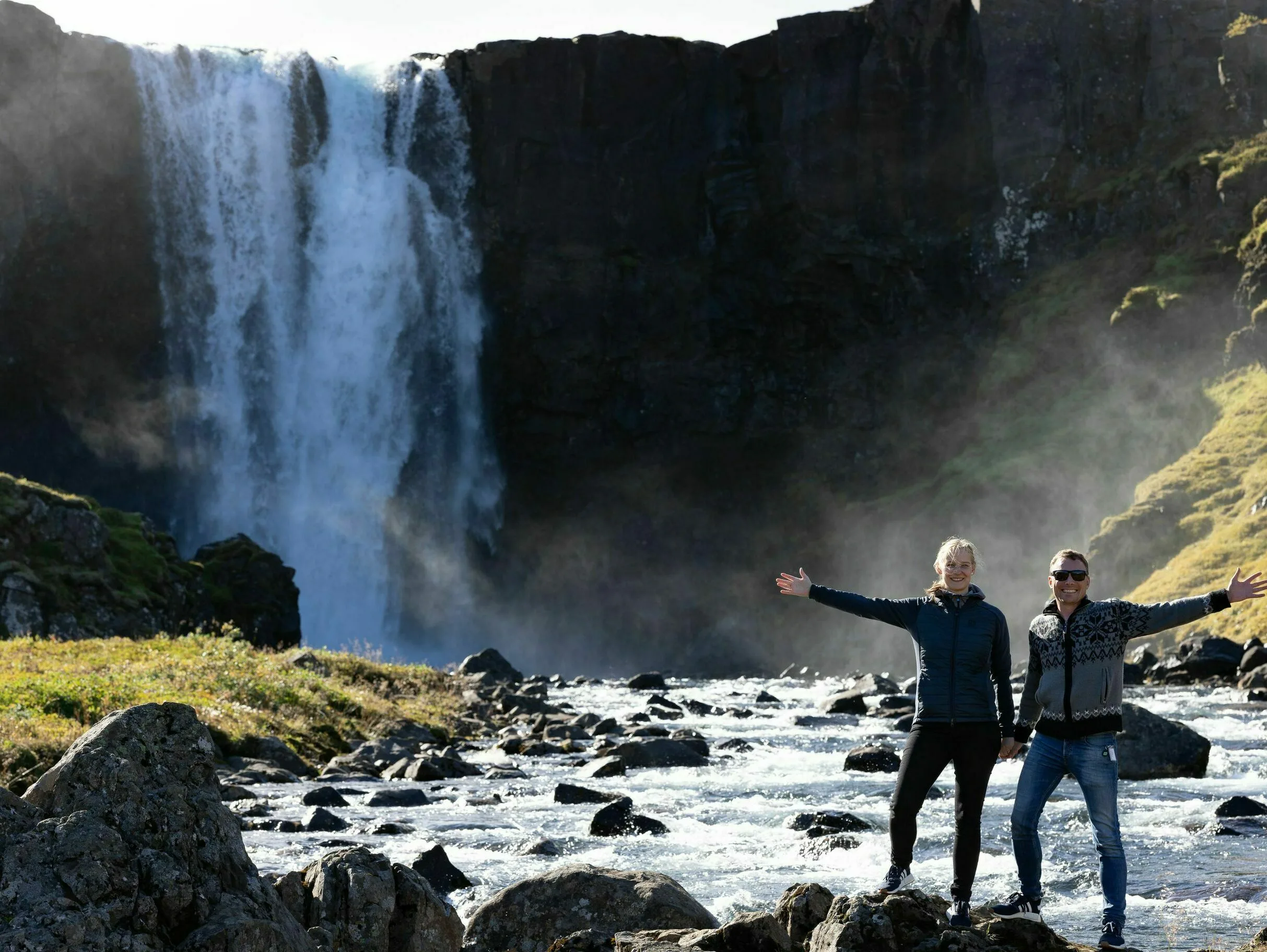 Ein Pärchen vor einem Wasserfall winkt fröhlich in die Kamera