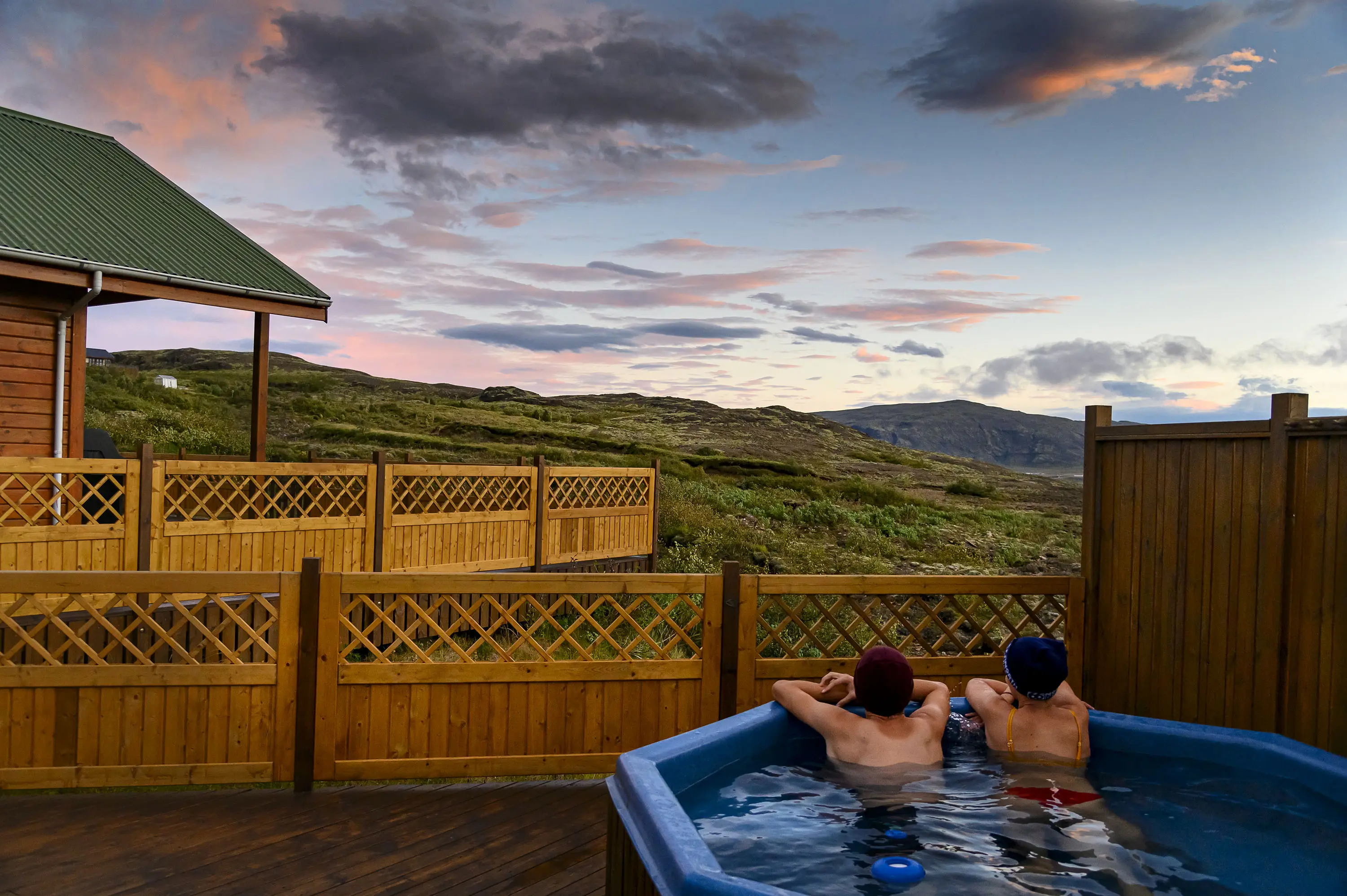 Island Ferienhaus mit Hot Pot auf der Terrasse, in dem 2 Personen den schönen Sonnenuntergang genießen
