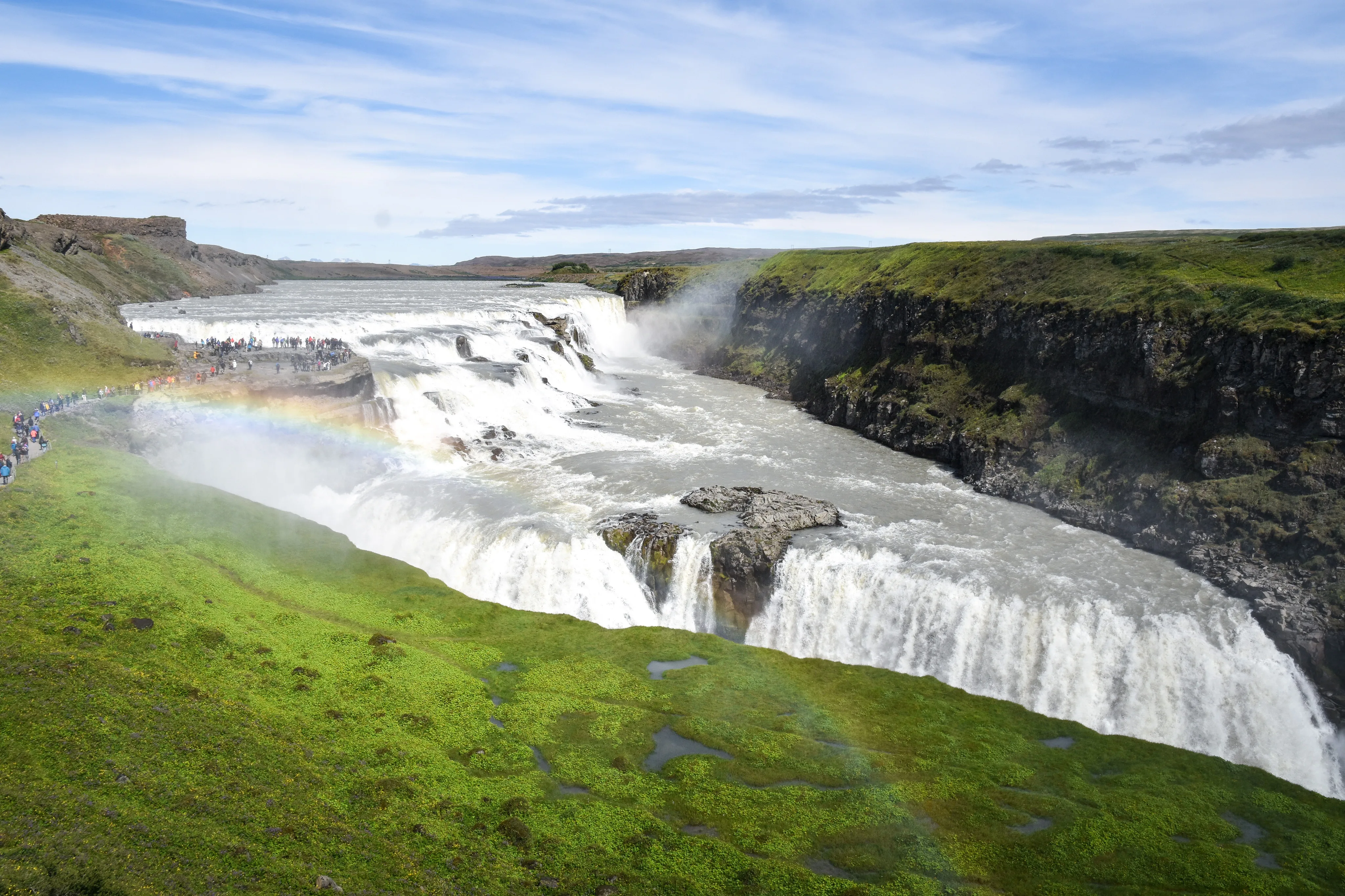 Luftaufnahme des imposanten Gullfoss Wasserfalls, über dem ein Regenbogen erscheint, daneben steht eine große Gruppe an Touristen, die den Wasserfall bewundert