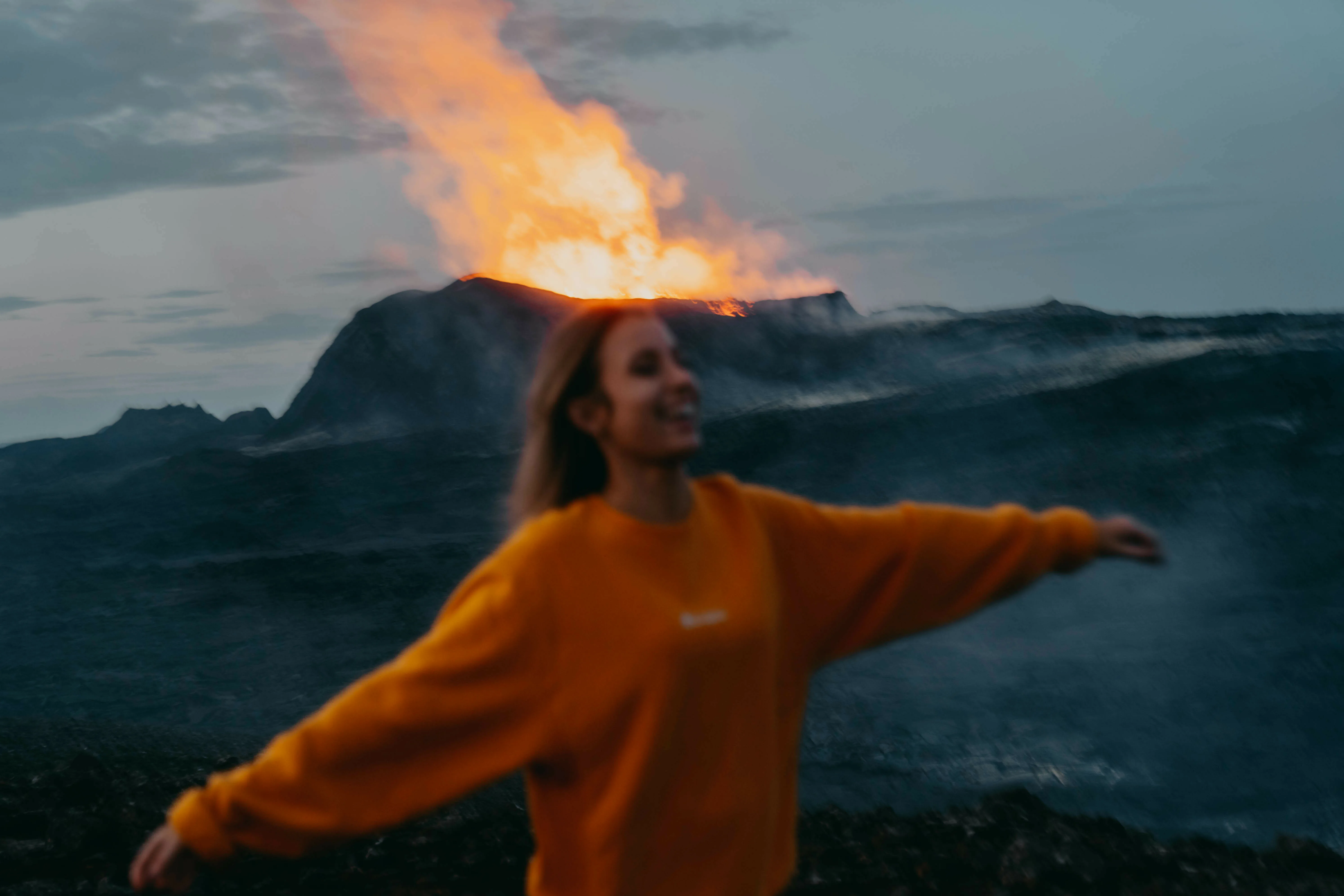 Island Vulkanausbruch; 
Frau in orangenem Pullover mit zur Seite ausgestreckten Armen steht lachend vor ausbrechendem Vulkan mit orangener Lava im Hintergrund. Leicht unscharf, um Bewegung der Frau zu sehen.