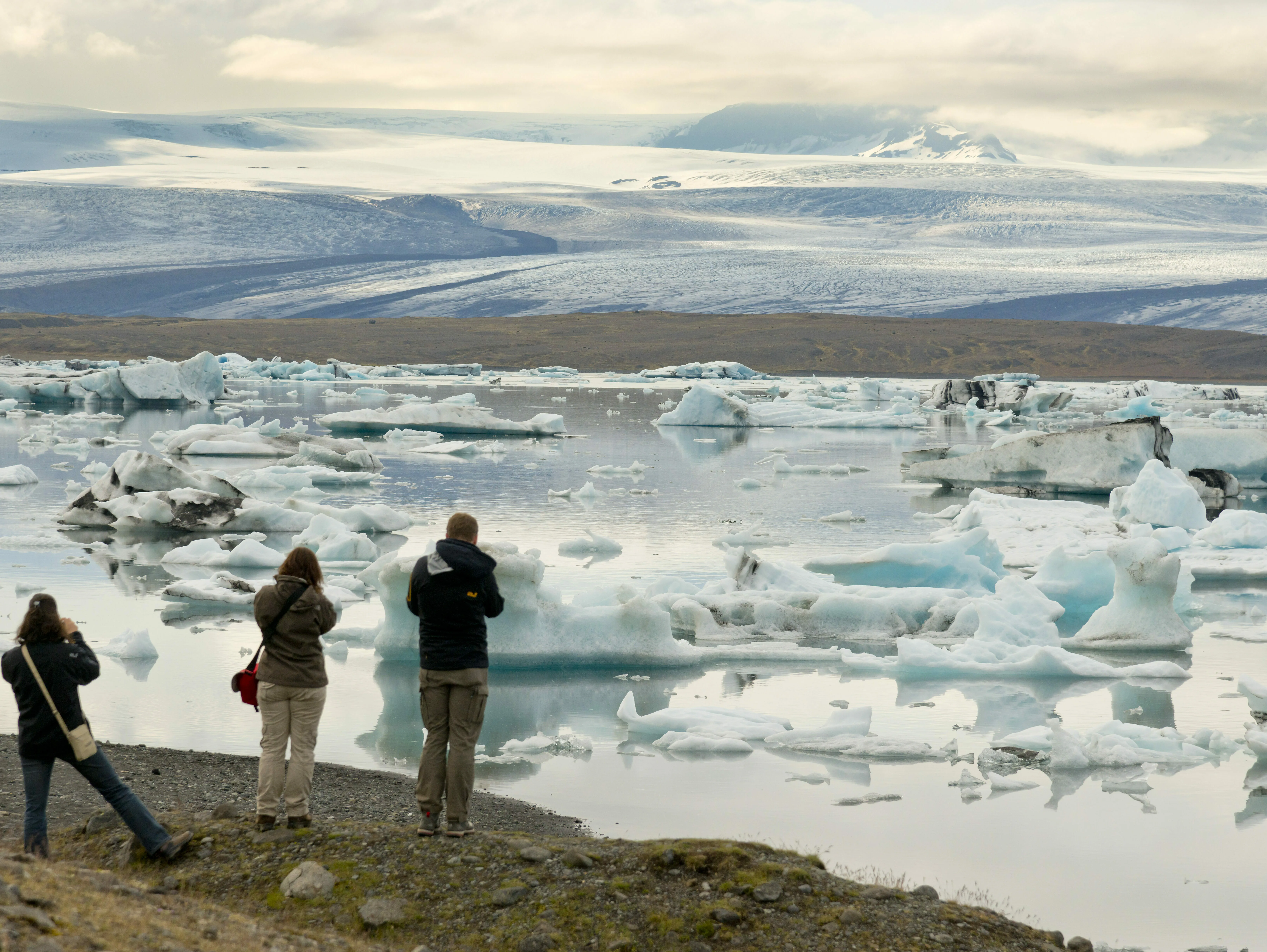 Drei Personen betrachten die Gletscherlagune Jökulsarlon und schießen Fotos. Die Lagune ist voll mit Eisbrocken. Im Hintergrund sieht man den Gletscher Vatnajökull.