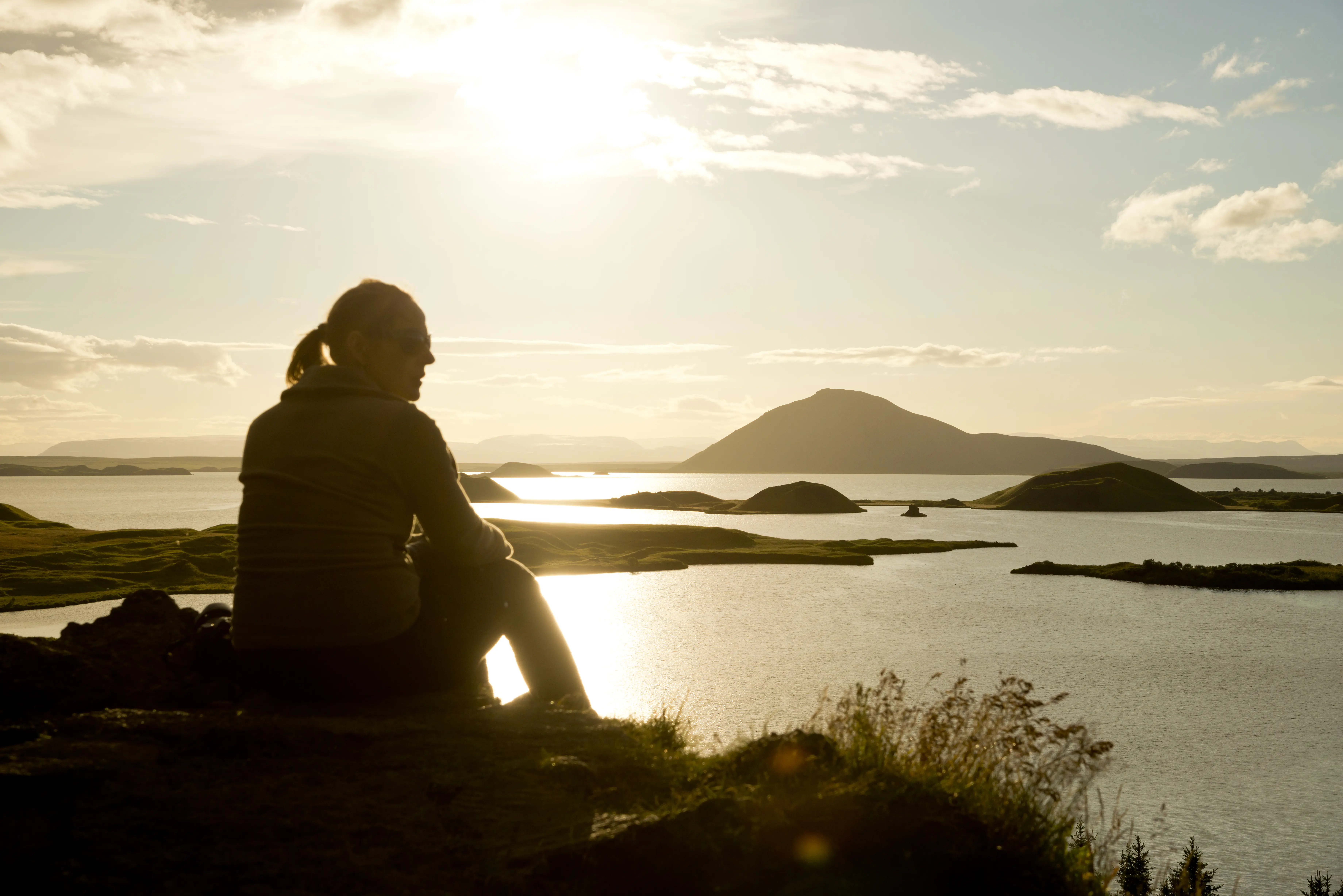Nordisland myvatn sonnenuntergang wandern foto thomas linkel Eine Person sitzt in der Abendsonne auf einem Hügel und blickt über den friedlichen Myvatn-See mit seinen kleinen Inseln und einem markanten Vulkan im Hintergrund.
