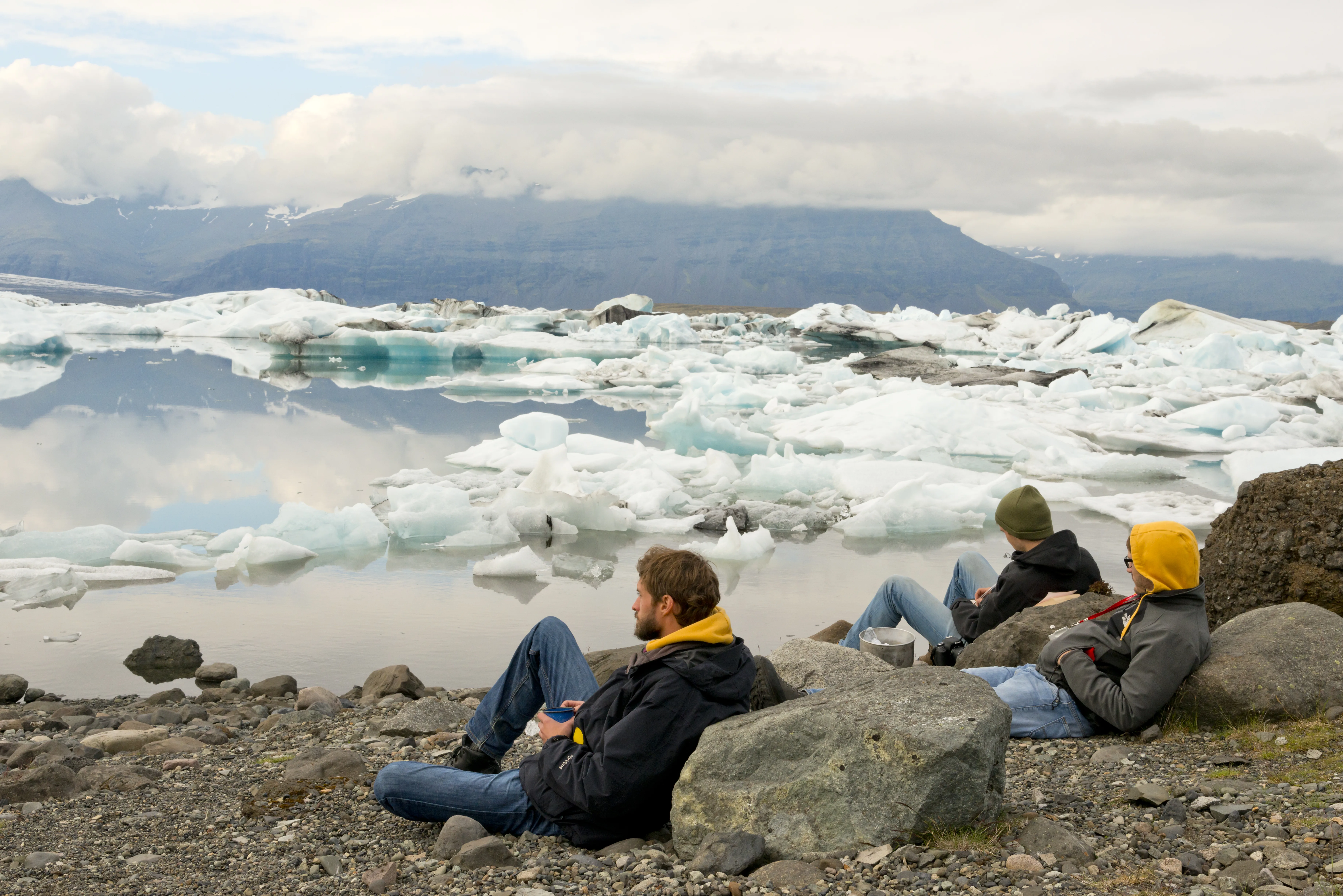 Südisland Gletscherlagune Jökulsarlon, Foto: Thomas Linkel
Drei Personen schauen an Felsen lehnend auf die Gletscherlagune Jökulsarlon, in der große Eisberge schwimmen. Im Hintergrund ist ein Berg mit Wolken verdeckt