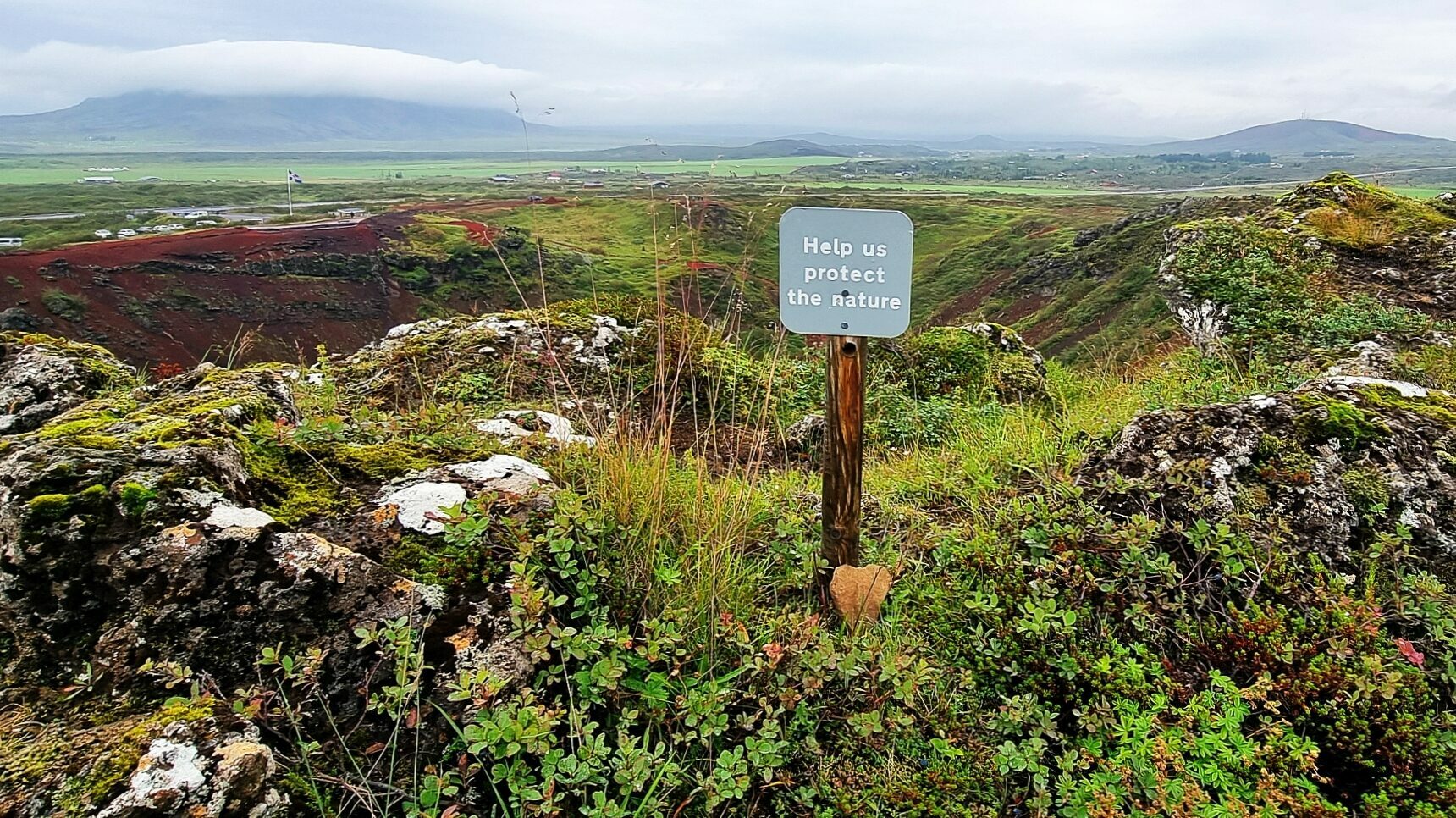 Island Nachhaltigkeit - Schild: help us protect the nature steht auf einem hügeligen Wandergebiet mit kleinen Sträuchern und Moosen. Im Hintergrund Straßen und Islandflagge erkennbar. Bewölkter Himmel.